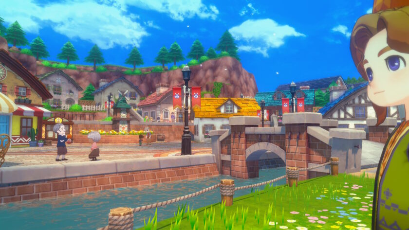 A screenshot from Story of Seasons: Grand Bazaar captures a vibrant town scene with cobbled streets, colourful houses, and a stone bridge crossing a gently flowing canal. In the foreground, a character with brown hair and a yellow-striped outfit stands to the right, partially facing the camera. Across the canal, two townsfolk chat near a flower stall, with banners and lampposts lining the street. The background features pine-covered cliffs under a bright blue sky with drifting petals, creating a cheerful and welcoming atmosphere. Published on: LadiesGamers.